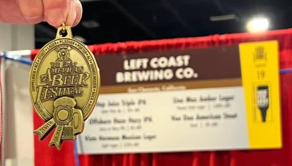 A person holds a gold medal from the Great American Beer Festival in Denver, Colorado. In the background is a booth for Left Coast Brewing.