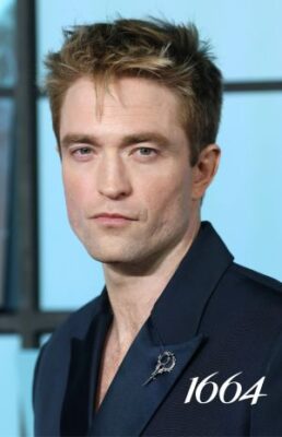 Robert Pattinson in a dark blue suit with a silver pin on the lapel, posing in front of a window.