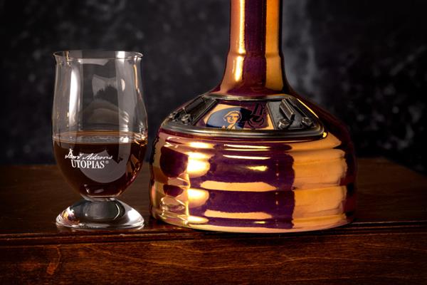 A close-up of a Samuel Adams Utopias beer bottle and a snifter glass filled with the dark, uncarbonated beer, sitting on a wooden surface.