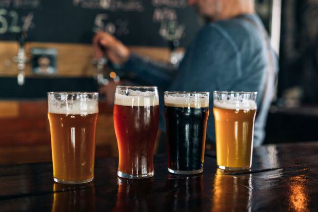 Four glasses of craft beer on a wooden bar counter showcasing different styles and colors, with a bartender pouring beer