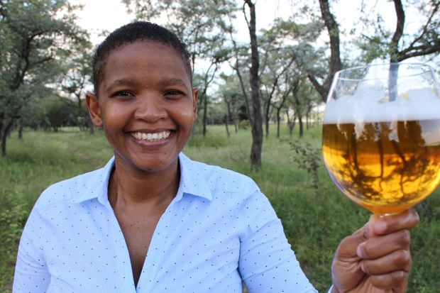 A smiling African woman holds up a large glass of beer outdoors in a natural, grassy landscape with scattered trees.