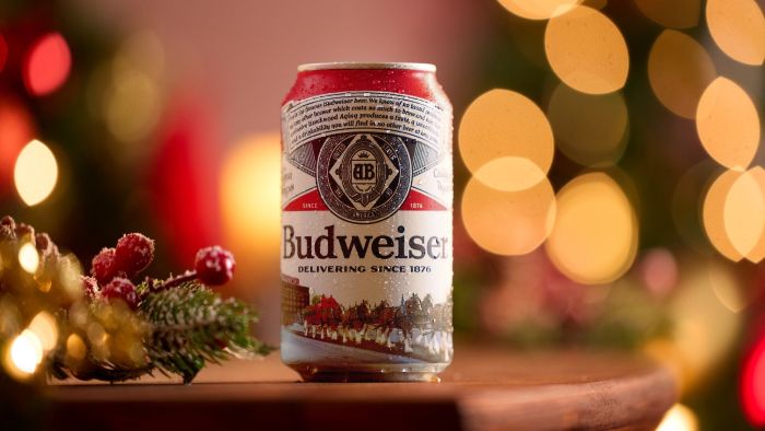 2025 Budweiser holiday can on a wooden table, in front of a holiday background with blurred Christmas lights and pine needles. The can's label features the Clydesdale horses pulling a sleigh in a snowy scene.