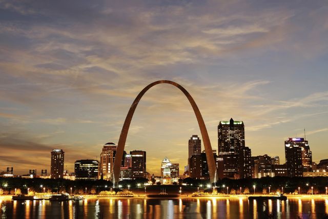 A stainless steel Gateway Arch stands illuminated against the St. Louis skyline at twilight, with the city lights reflecting on the Mississippi Rive