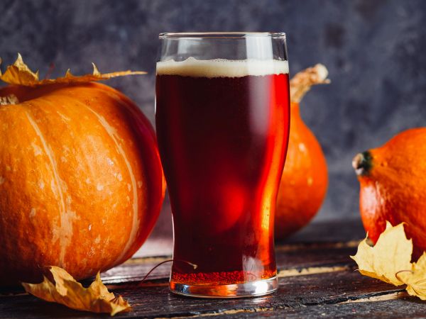 A glass of pumpkin beer surrounded by pumpkins and autumn leaves on a rustic wooden table.