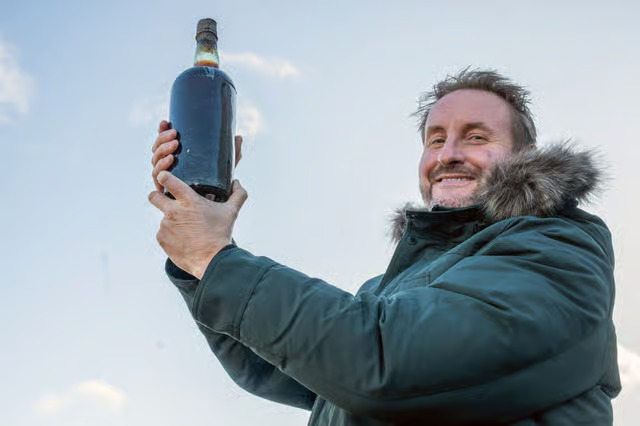Dougal Gunn Sharp  smiling in a green, fur-trimmed jacket as he holds up historic Arctic ale.