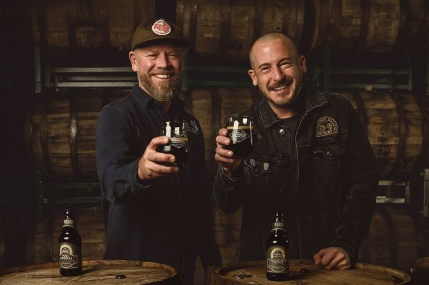 Two smiling men raise glasses of dark beer in a cellar full of aging wooden barrels, with two branded beer bottles standing on barrels in the foreground