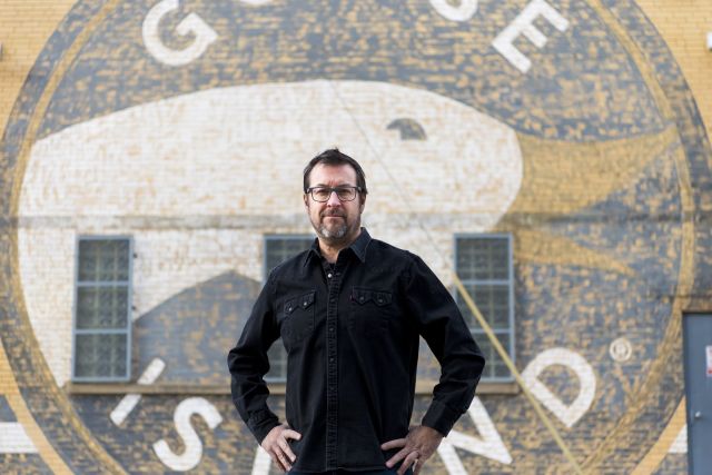 Todd Ahsmann with dark hair and glasses wearing a black button-down shirt stands centered in front of a large circular wall mural featuring a goose logo