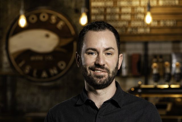 John Zadlo smiles at the camera in an establishment with a blurred 'Goose Island' logo in the background.