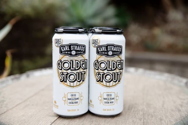 Two white cans of Karl Strauss Golden Stout beer sit on a wooden surface outdoors.
