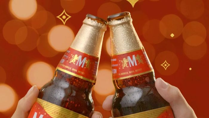 Two hands holding and clinking two bottles of Modelo beer against a warm red background with festive lights