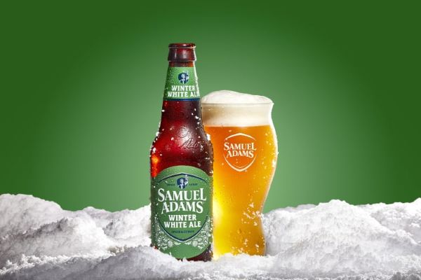 A bottle and a glass of Samuel Adams Winter White Ale beer sitting in a bed of snow, against a green background