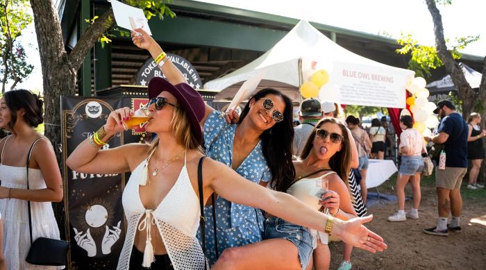 Three young women having fun at an outdoor beer festival, with tents and other people in the background.