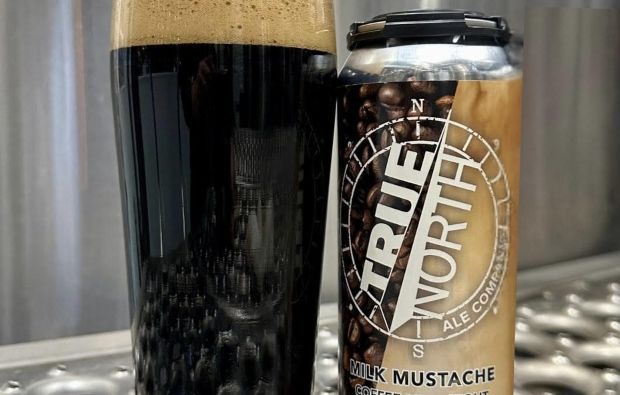 A glass of dark stout beer with a frothy head next to a can of True North Ale Company Milk Mustache Coffee Milk Stout, both resting on a metallic surface.