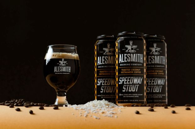 AleSmith Speedway Stout beer cans and glass, with coffee beans and toasted coconut flakes on a table.