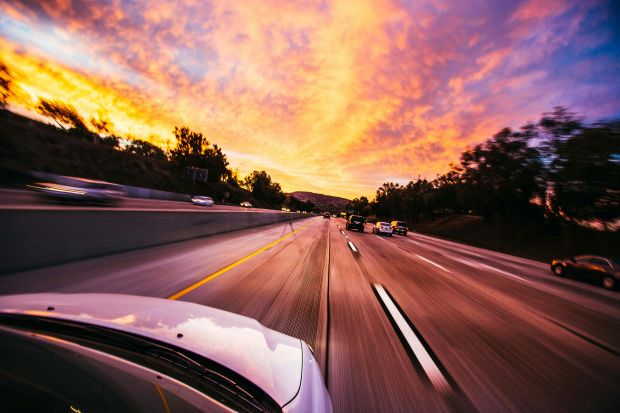 A point-of-view pespective from a moving car on a multi-lane highway, with other cars in motion, at sunset, featuring vibrant orange, yellow, and purple cloud