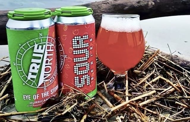 Two cans of True North Ale Co. 'Eye of the Storm' Hurricane Sour and a poured glass of the beer on a beach.