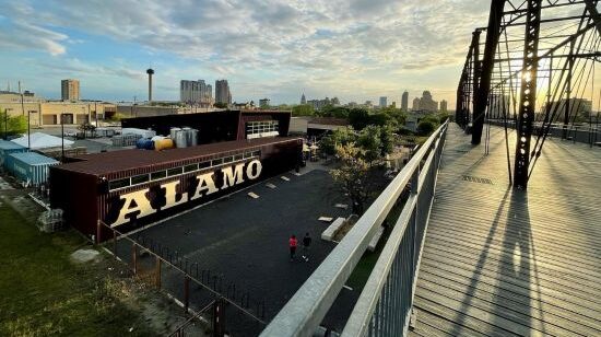 A view from a metal truss bridge of the Alamo Beer Company building in San Antonio at sunset, with the city skyline in the background.