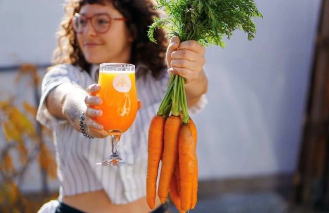A person smiling and holding up a glass of bright orange Rtio Beerworks "King of Carrot Flowers" Saison beer next to a bunch of fresh carrots
