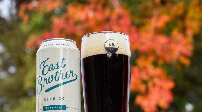A can and glass of East Brother Beer Co. Munich Dunkel, a seasonal dark lager against a a fall leaves backdrop.