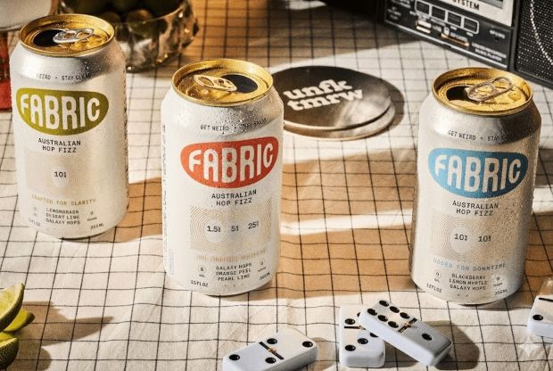Three chilled cans of Fabric Australian Hop Fizz in green, orange, and blue varieties arranged on a checkered table with dominoes.