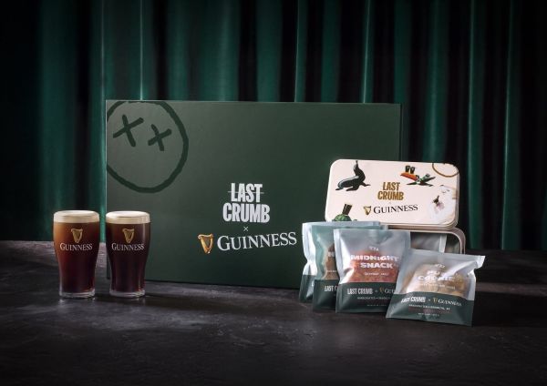 Two glasses of Guinness stout beer, packaged cookies in pouches and a tin, and a large green gift box with the "Last Crumb x Guinness" logo, set against a dark green curtain background.