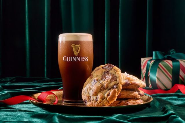 A glass of Guinness next to a stack of chocolate chip cookies on a gold tray, with a festive wrapped gift in the background.