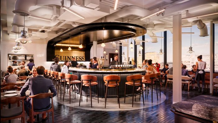 A lively, modern bar and restaurant area within the Guinness Open Gate Brewery in Covent Garden, London, featuring a central oval-shaped bar, bar stools, and seated diners with large windows in the background