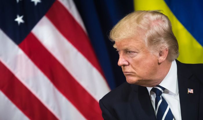 President Donald Trump in profile, wearing a suit with a U.S. flag pin, seated in front of the American flag and a blue and yellow flag.