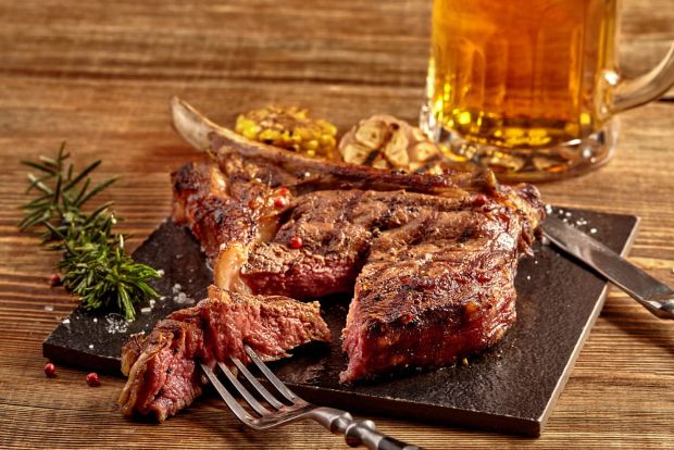 A large, cooked bone-in steak, which has been sliced and is presented on a dark serving board with a fork. A full glass of beer in a mug is visible in the background, all set on a rustic wooden table.