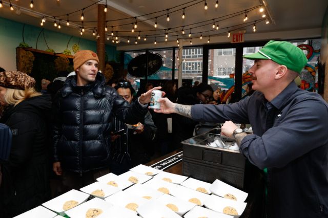 A man in a green cap and black shirt hands a drink to a man in an orange beanie and black puffer jacket, at Dogfish Head pop-up event in NYC.