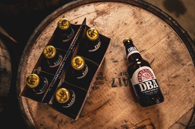 A six-pack of Firestone Walker DBA beer bottles, with one bottle standing alone, all resting on a large wooden barrel in a dimly lit setting