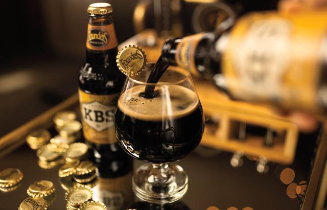 A bottle of Founders Kentucky Breakfast Stout (KBS), being poured into a snifter glass, with a full bottle and several bottle caps nearby on a wooden counter.