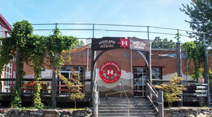 he exterior entrance of the Highland Brewing Company in Romeville, NC on a sunny day. The building has a large central entrance with stairs leading up to it and decks on either side with tables and chairs.