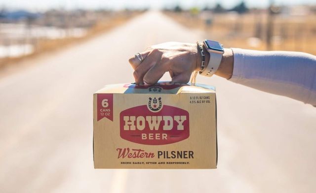 A hand holds a six-pack of Howdy Beer Western Pilsner while standing on an empty, open road.