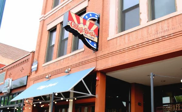 The exterior facade of Oskar Blues Grill & Brew, a restaurant located in a historic orange-brick building in downtown Colorado Springs, with a large branded sign and a blue awning over the patio