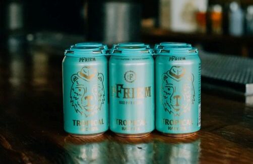 Six-pack of pFriem Tropical Hazy IPA beer cans sitting on a wooden bar top.