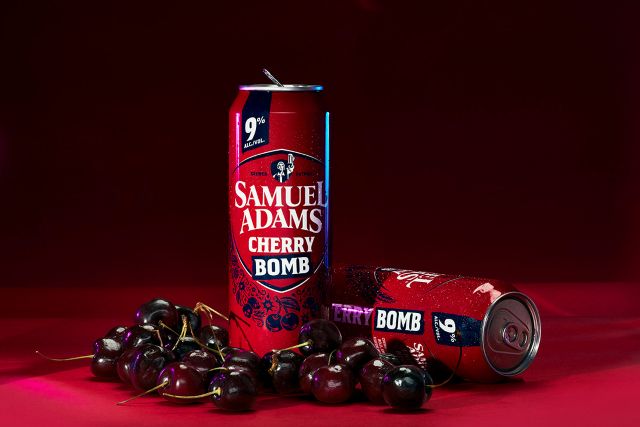 Two cans of Samuel Adams Cherry Bomb beer and a pile of fresh cherries on a red surface.