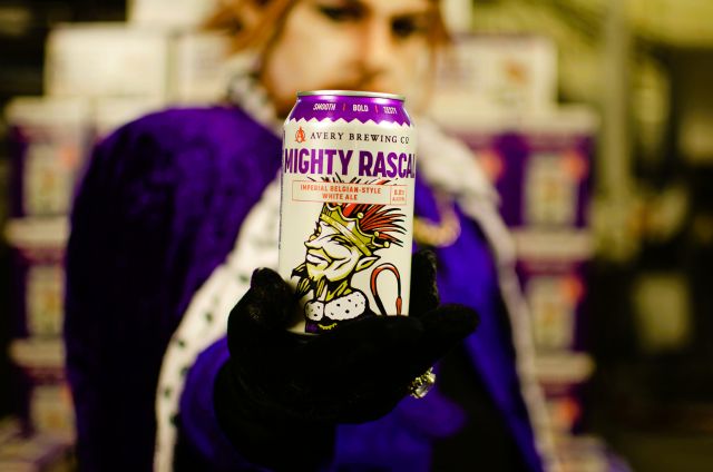 A person in a royal purple costume holding up a 12oz can of Avery Brewing Co. Mighty Rascal, an Imperial Belgian-Style White Ale