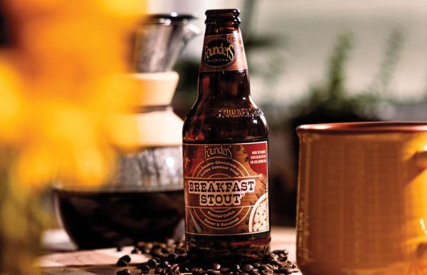 A bottle of Founders Breakfast Stout beer sits among scattered coffee beans, a glass coffee brewer, and a mug
