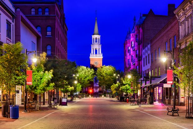 Night view of Church Street Marketplace in Burlington, Vermont, featuring a long pedestrian brick-paved street lined with shops and a glowing white church steeple in the distance."