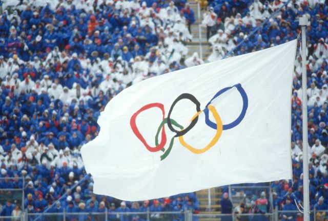 A white Olympic flag featuring five interlocking rings (blue, yellow, black, green, and red) flying at a stadium filled with spectators in blue and white attire.