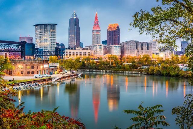 The Cleveland, Ohio skyline at dusk, with the city lights beginning to glow and reflecting in the tranquil water of the Cuyahoga River in the foreground.