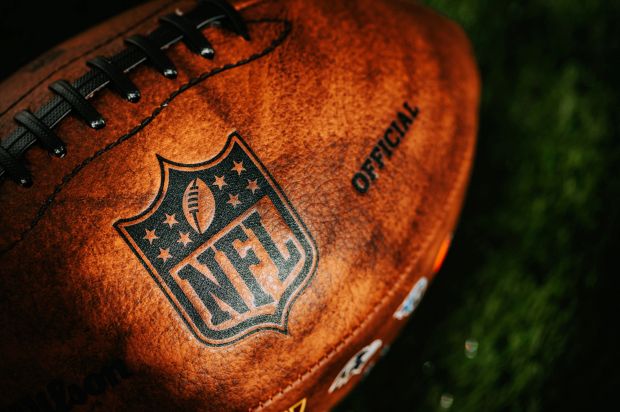 A close-up shot of a brown, textured American football with the official NFL shield logo stamped on its side, resting on green turf.