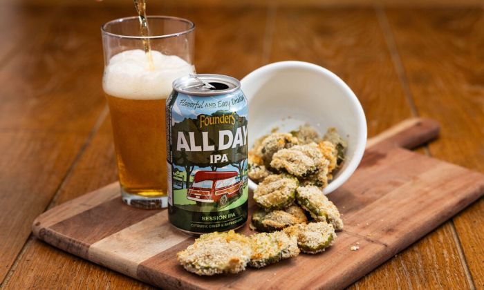 A can of Founders All Day IPA sits on a wooden board next to a glass of poured beer and a bowl of breaded, fried pickle