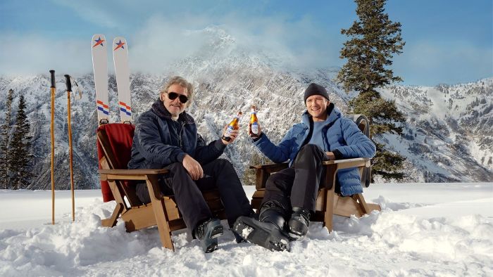 Kurt Russell and Lewis Pullman, both in ski gear, are sitting in wooden chairs on a snowy mountain and toasting with Michelob Ultra beers. This image is from a 2026 Michelob Ultra Super Bowl commercial called "The ULTRA Instructor