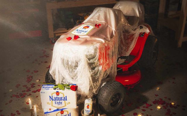A red riding mower draped in a sheer Lingerie-like cover with rose petals and lit candles, and a large box of Natural Light beer in the foreground.
