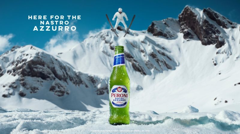 A green glass bottle of Peroni Nastro Azzurro beer sitting in snow with a skier jumping in the snowy mountains in the background