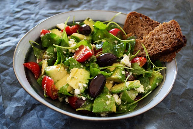 A bowl of salad with avocado, tomatoes, feta cheese, olives, and mixed greens, served with two slices of dark whole-grain bread.