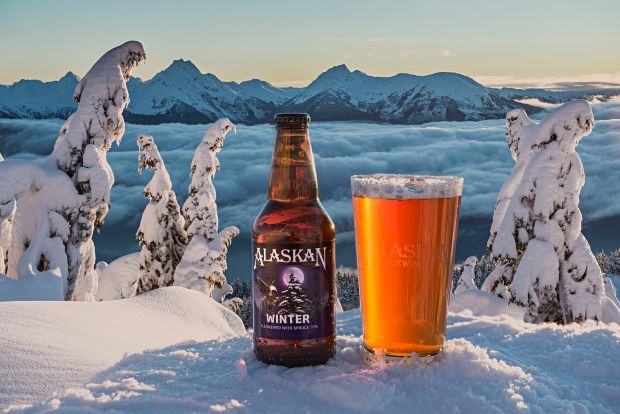 A bottle and a pint glass of Alaskan Winter Ale sit in the snow, overlooking a vast, cloud-filled valley and jagged, snow-capped mountain peaks at sunset.