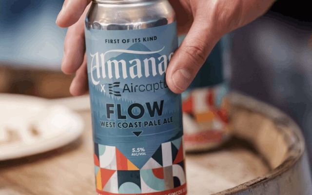 A and holding a blue can of Almanac x Aircapture Flow West Coast Pale Ale, featuring colorful geometric patterns at the bottom and a 5.5% ABV label.